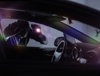 A hooded car thief shines a flashlight into a car at night.