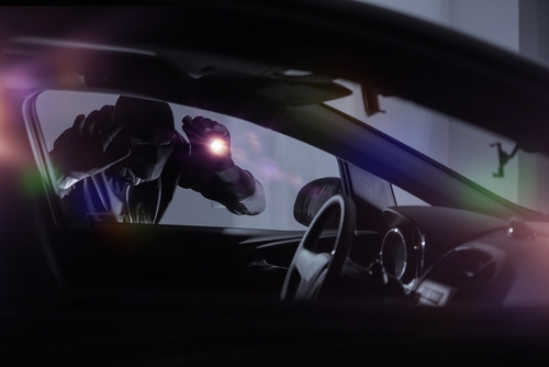 A hooded car thief shines a flashlight into a car at night.
