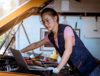 woman looking at a laptop while working on a car engine