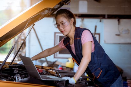 woman looking at a laptop while working on a car engine